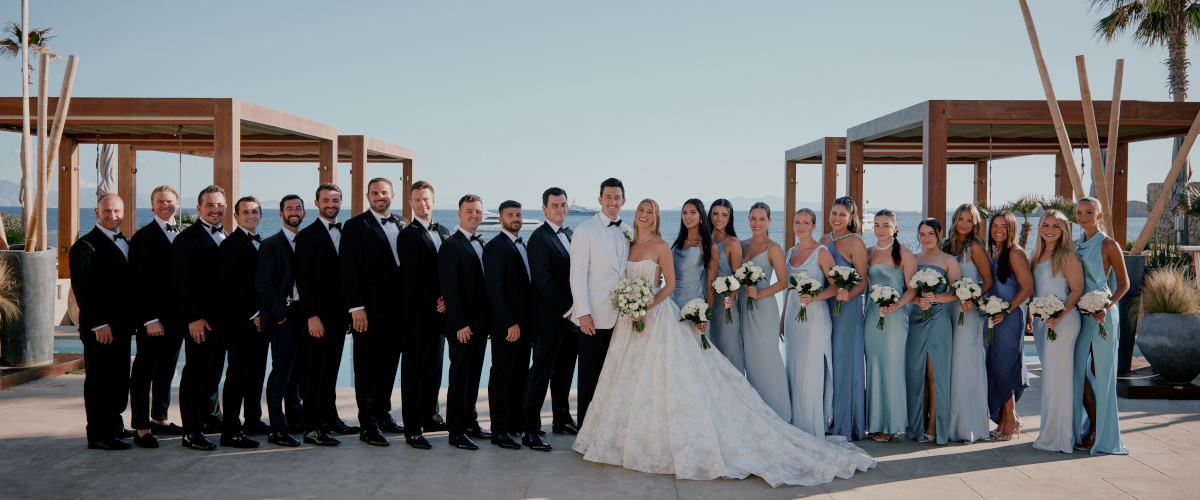 Luxury wedding in Mykonos, bride and groom posing with bridal party with Formal attire, at the poolside of a luxury hotel wedding venue overlooking the Aegean Sea.