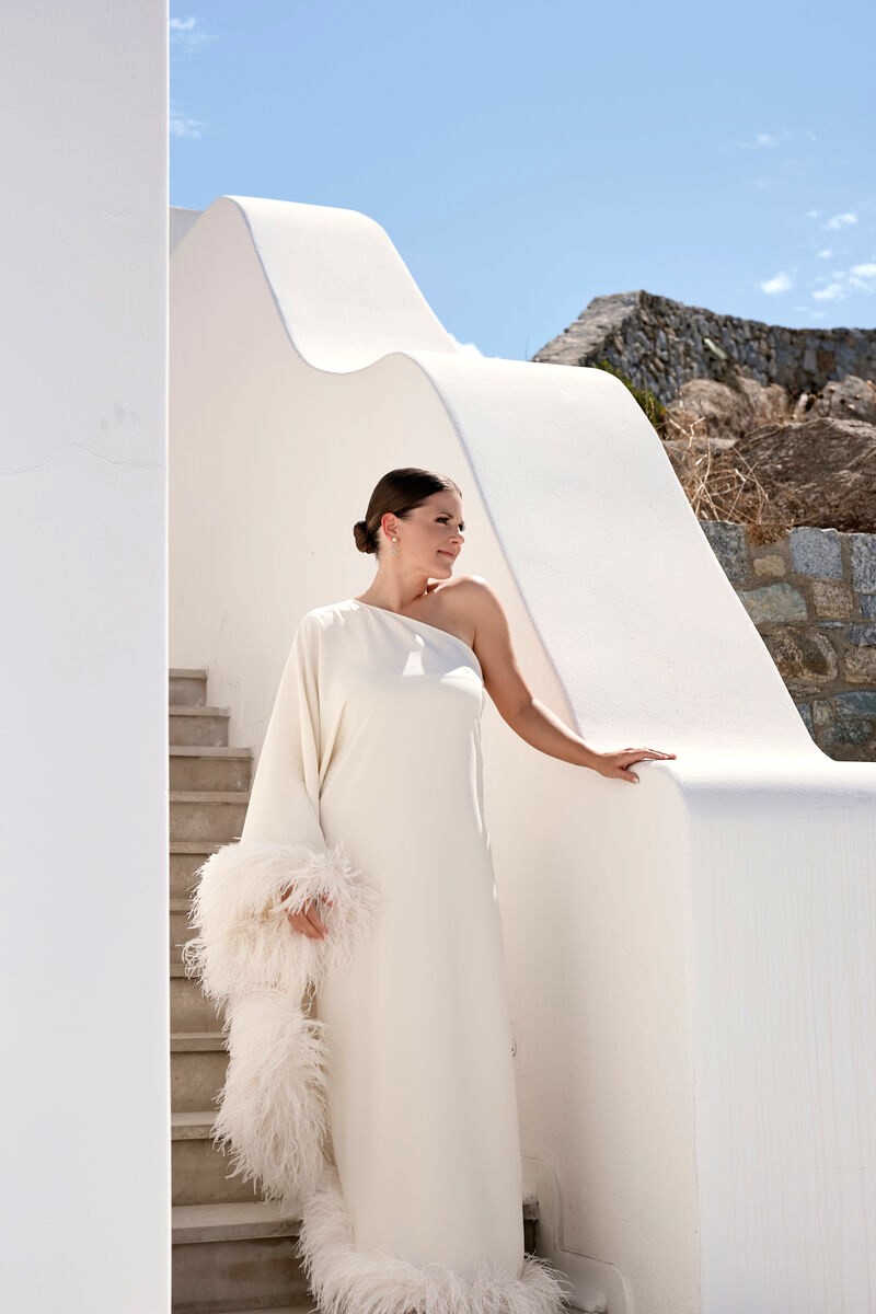 Bride in an elegant one-shoulder gown with feather details standing on white stairs at a boutique hotel in Mykonos during an intimate luxury wedding elopement