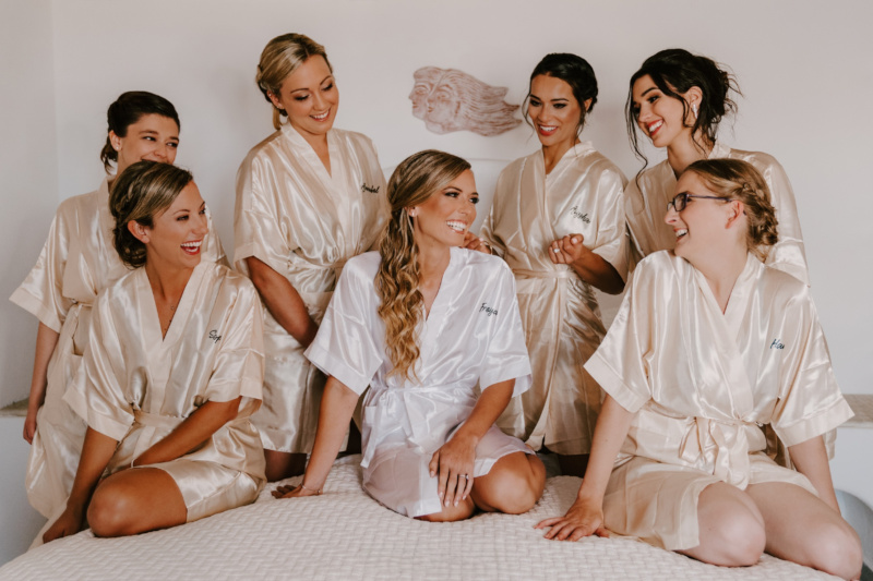 Bride's wedding party in Mykonos, bridesmaids sitting on a bed wearing matching satin robes. Hair and make up stylists pampered them.